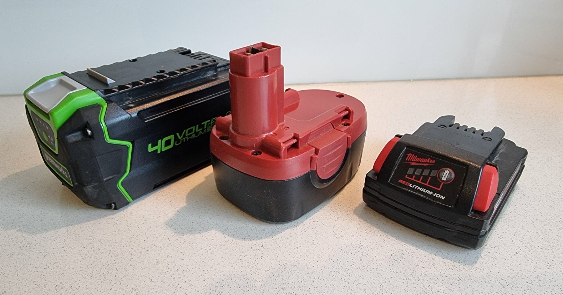 Three different types of rechargeable power tool batteries are displayed on a light countertop: a green 40-volt, a red rectangular, and a black Milwaukee lithium-ion battery.