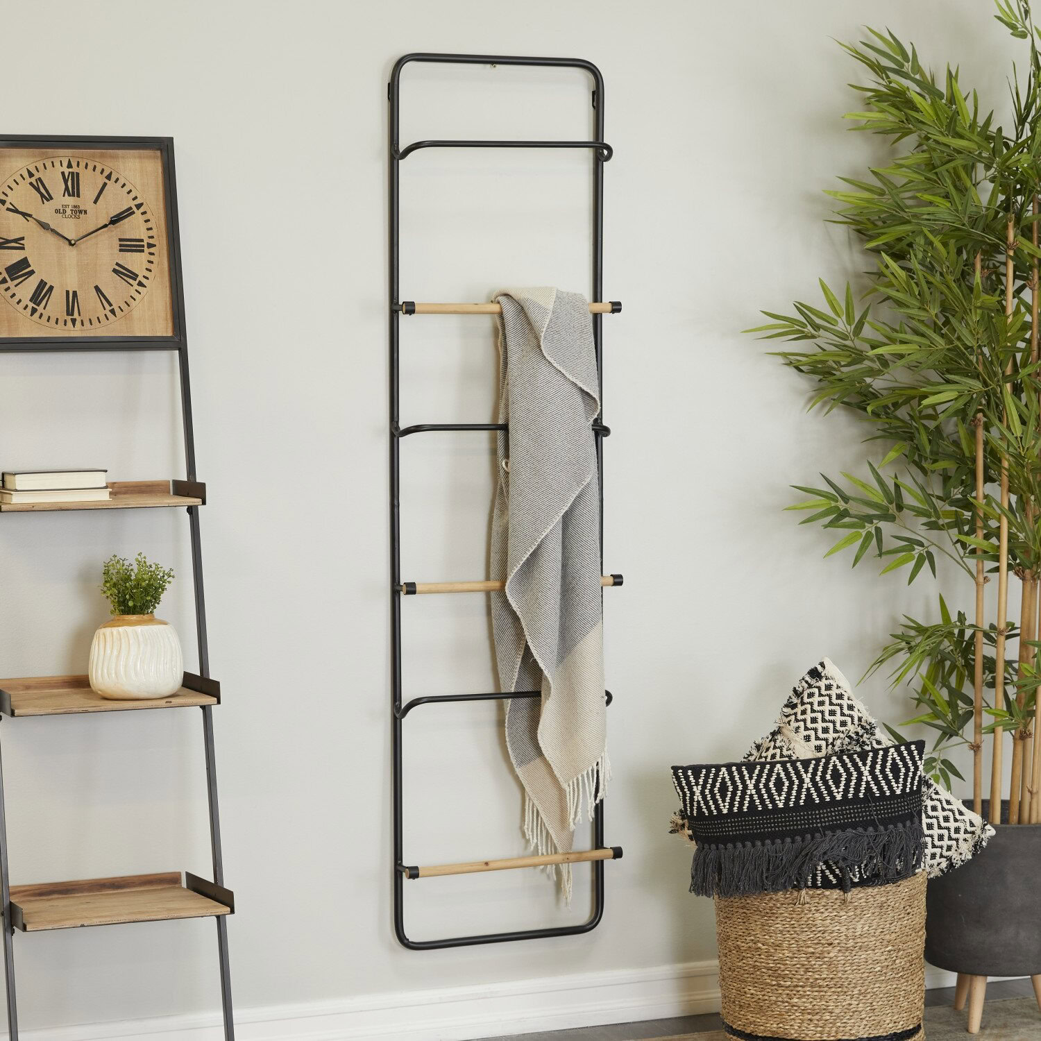 A metal ladder with wooden rungs leans against a wall, draped with a gray blanket. Nearby are a decorative clock, a shelf with books, and potted plants.