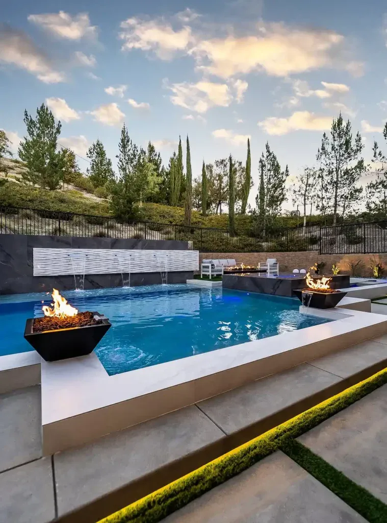 Modern outdoor swimming pool with two fire pits, water fountains, and landscaped surroundings, set against a backdrop of trees and a cloudy sky.