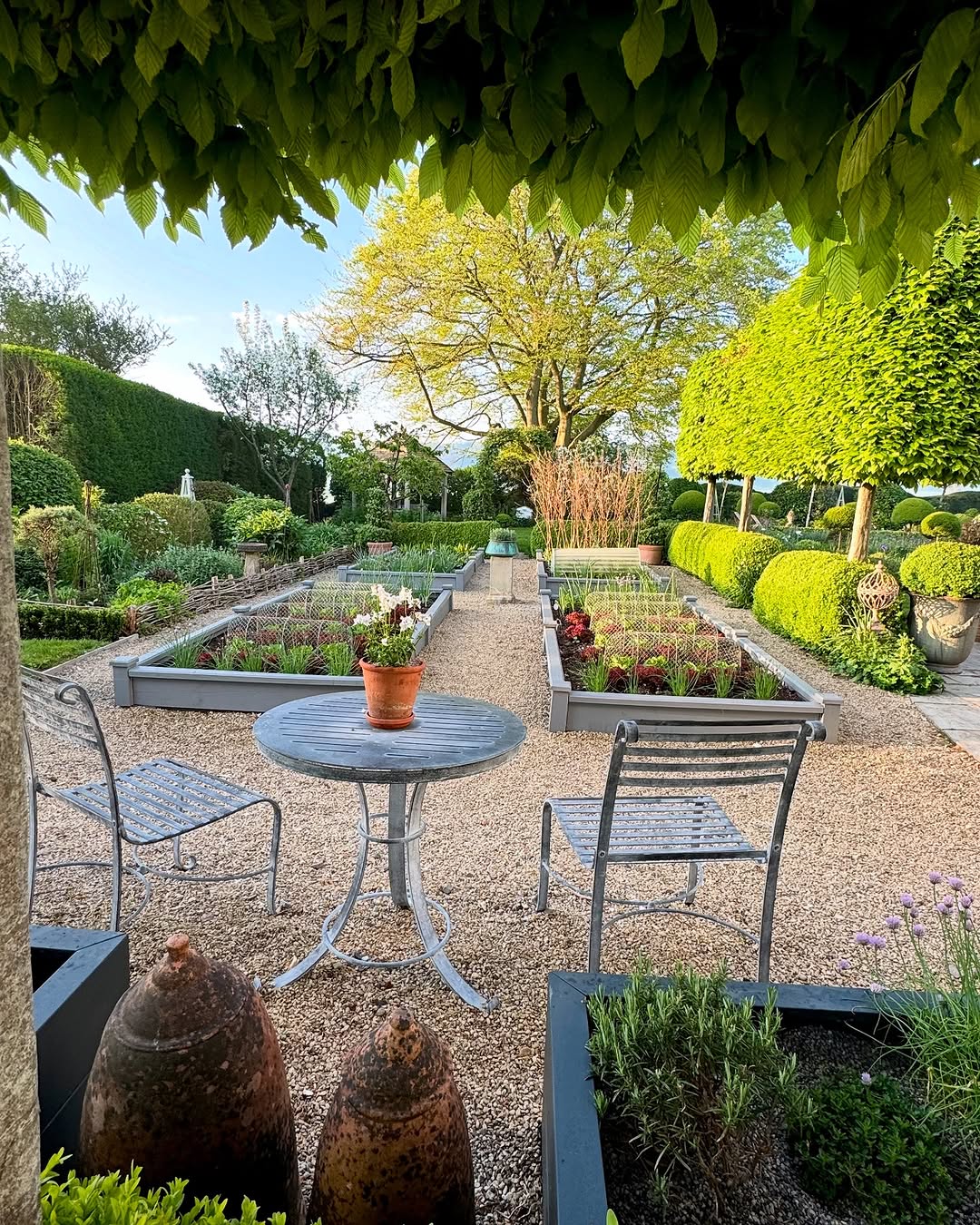 A gravel garden path with raised beds and trimmed hedges, viewed from a small table with two metal chairs and a potted plant in the foreground.