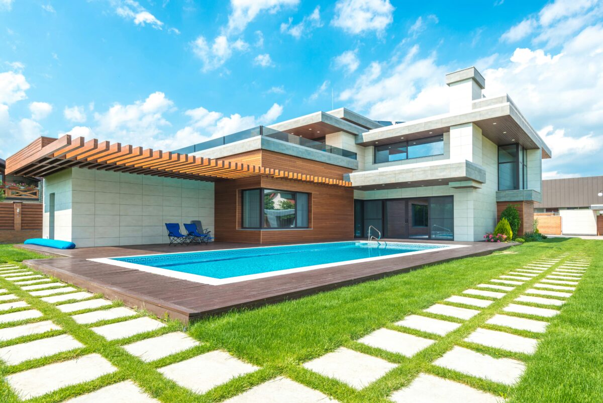 Modern two-story house with a swimming pool, wooden deck, pergola, and green lawn under a bright blue sky with clouds.