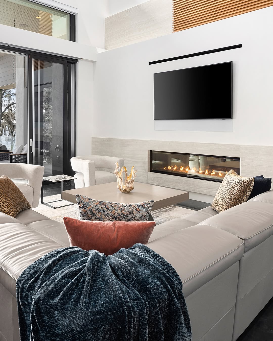 A modern living room features a sectional sofa, throw blanket, coffee table, decorative items, a linear fireplace, wall-mounted TV, and large windows with a view outside.