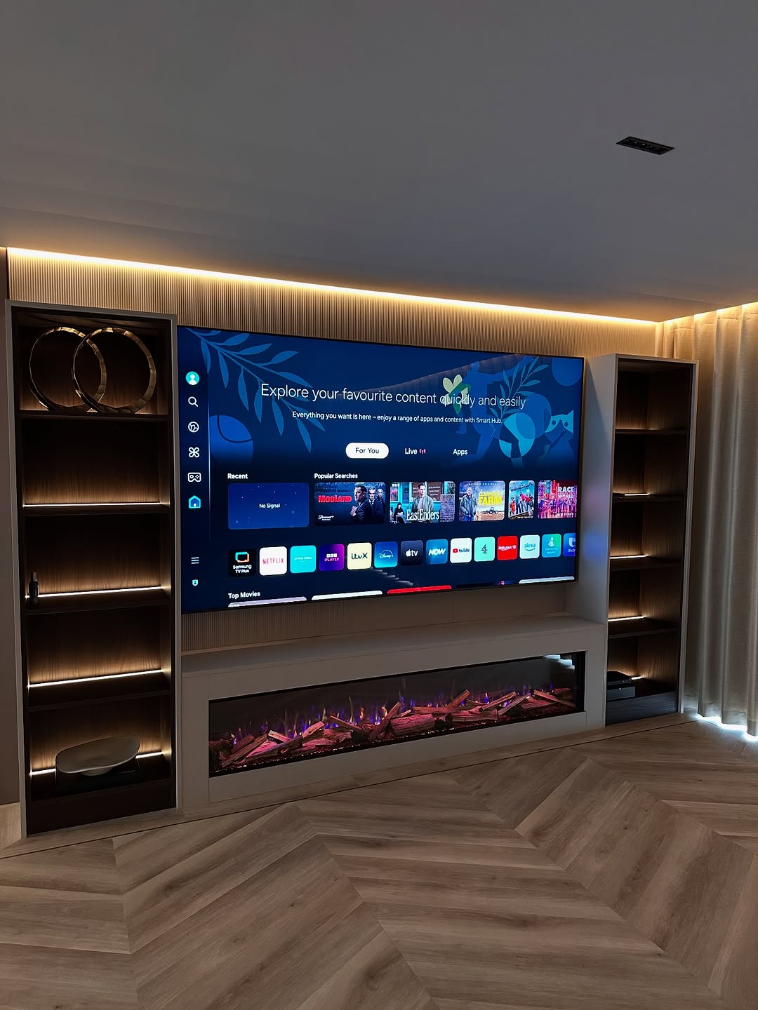 A large flat-screen TV is mounted on a wall with built-in shelves and an electric fireplace below. The TV displays a smart menu interface with various apps and streaming options.