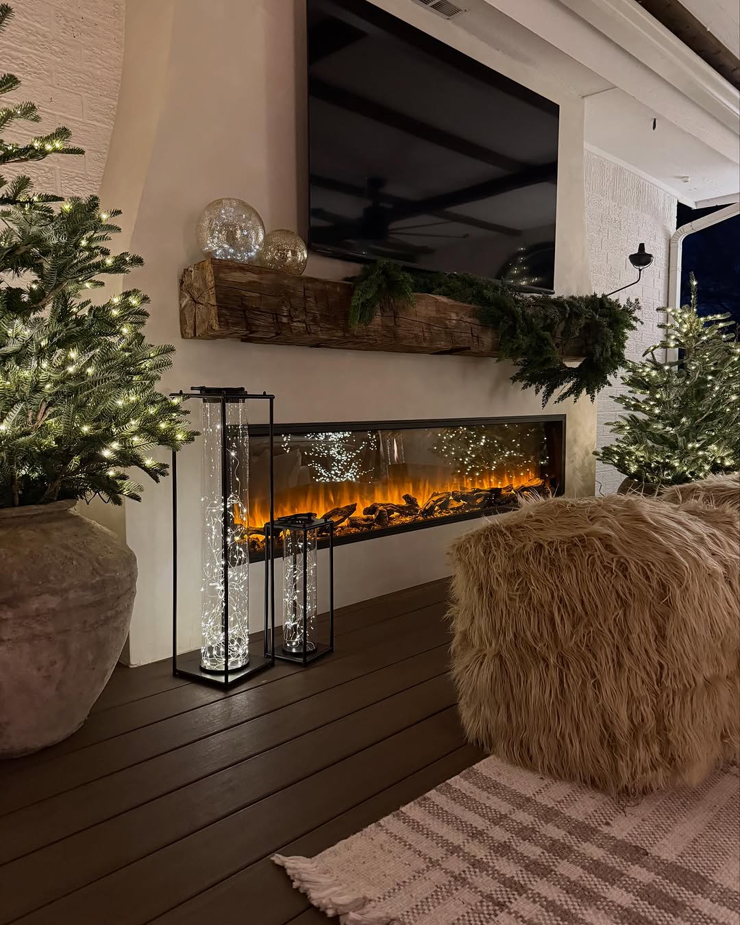 A modern living room with a lit electric fireplace, decorated mantel, lit Christmas trees, shaggy pouf, patterned rug, and glass cylinder lights.