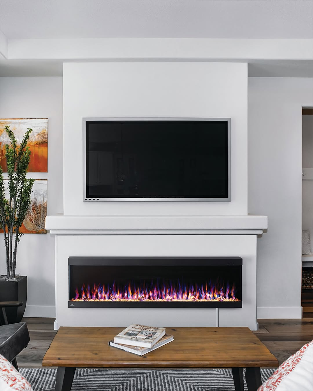 A modern living room with a wall-mounted TV above an electric fireplace, a wooden coffee table, and decorative art and plants on the side.