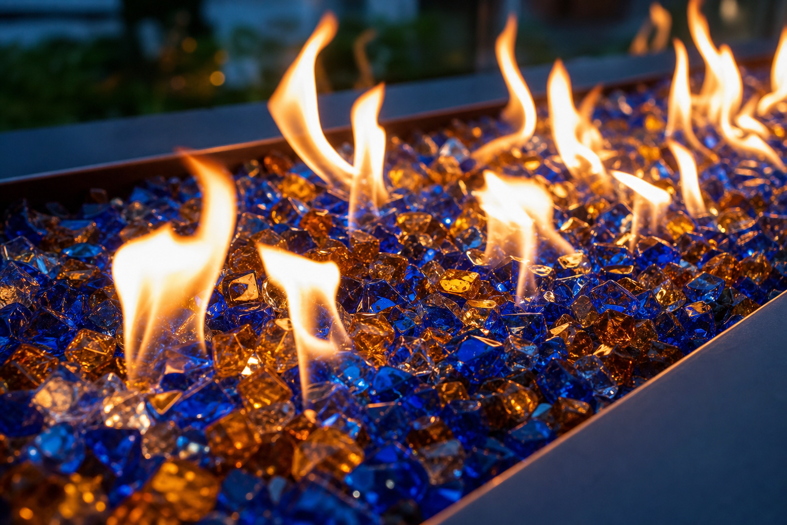 Close-up of blue and amber fire glass stones in a rectangular fire pit with yellow flames burning above them.