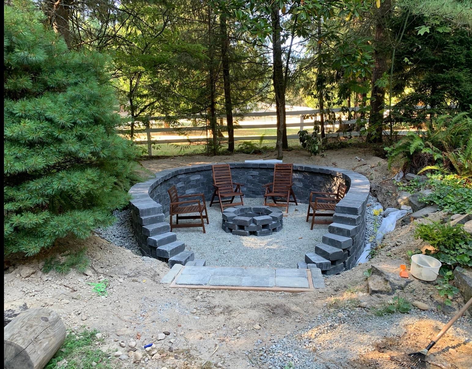 Finished sunken fire pit with curved block retaining wall, gravel floor and wooden chairs in a forest clearing