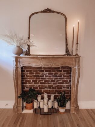Carved wood mantel against a wall with faux brick, pillar candles and a gilt mirror